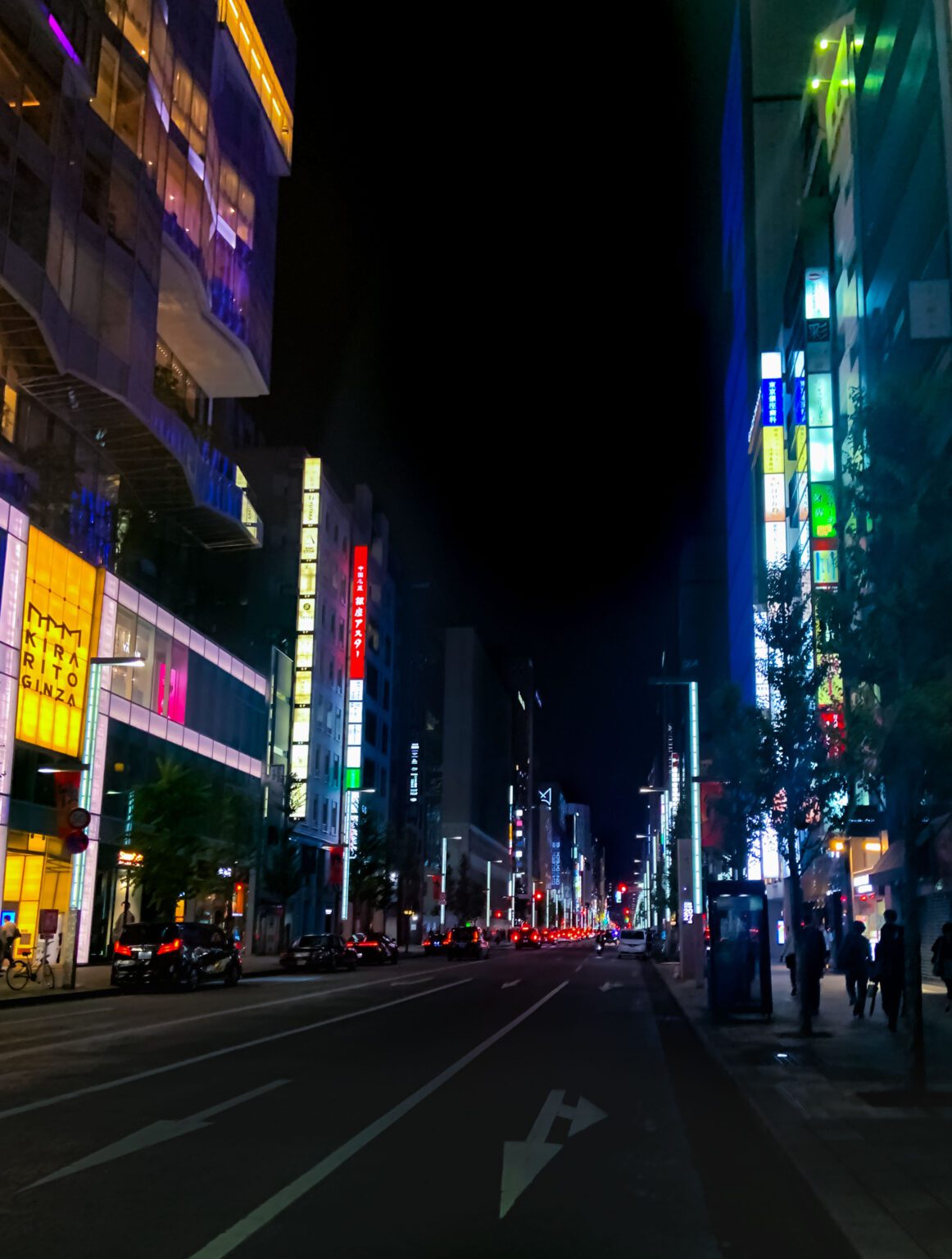 Main image of Ginza shopping district at night.Tall skyscapers exude neon lights as people walk beneath them. 7 must-go places in ginza.